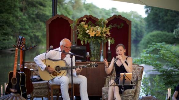 Ein Mann und eine Frau sitzen unter einem Dach im Freien, im Hintergrund ist Wasser zu sehen. Sei singt und er spielt Gitarre.