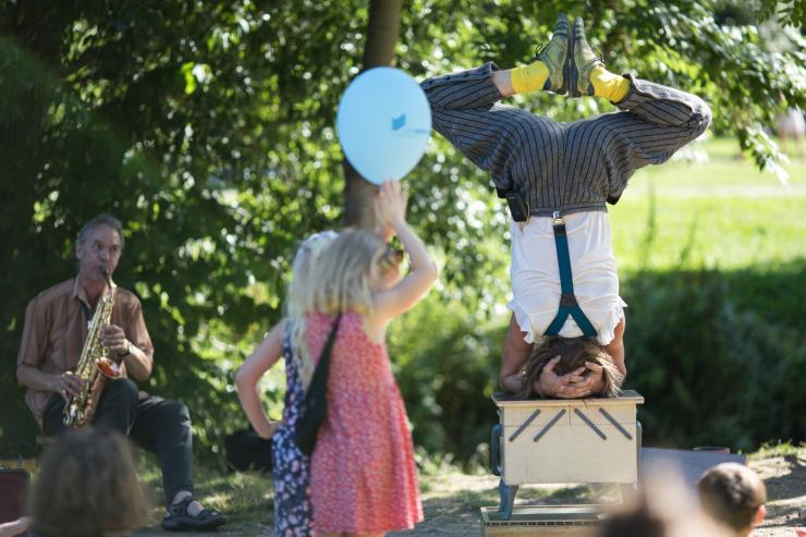 Artist balanciert im Kopfstand auf einer Kiste, danebben spielt ein Musiker Saxofon, vor ihnen Kinder mit Luftballons